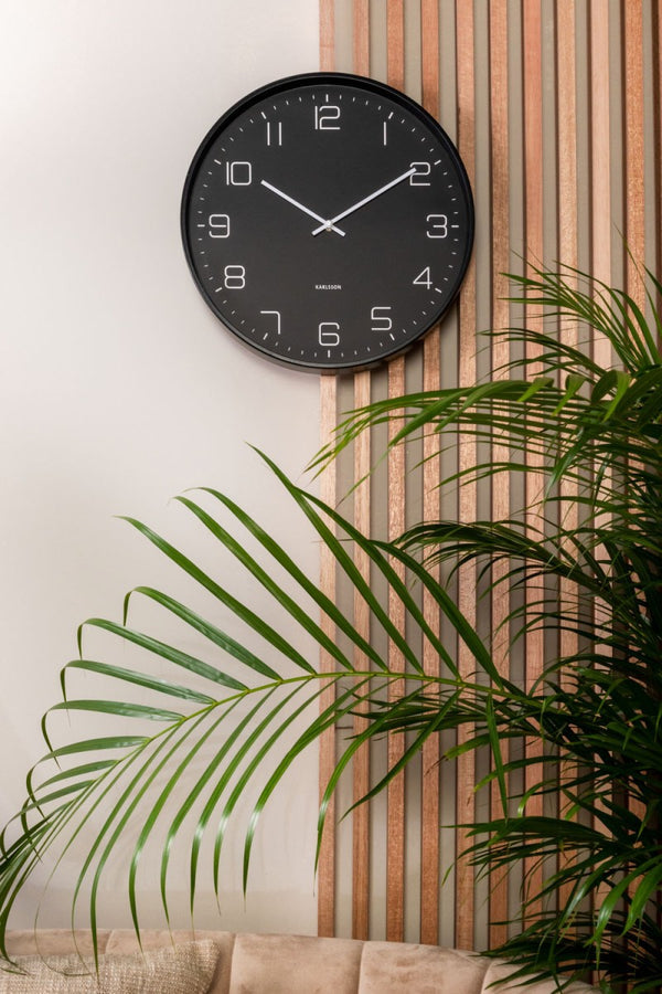 Karlsson Lofty Wall Clock in matte black on a timber slat wall beside a palm