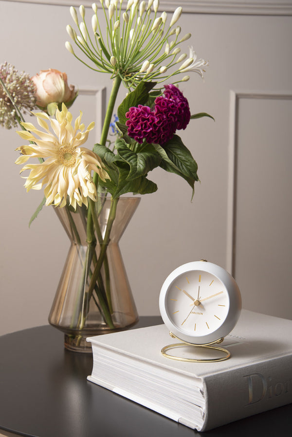 White Globe alarm clock on a stack of books beside a vase of flowers on a dark sideboard