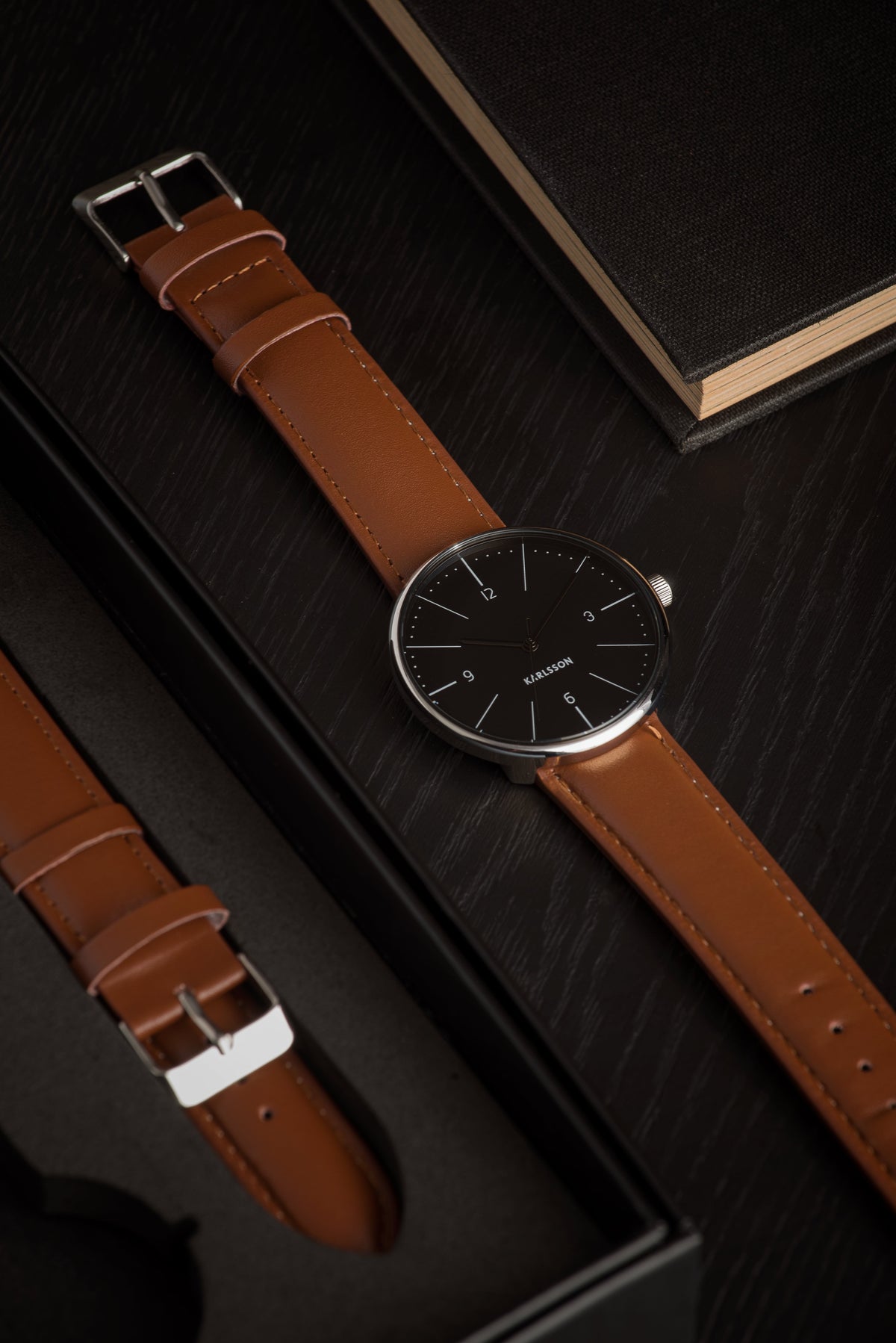 Karlsson Normann watch with a black dial on a dark surface beside a spare tan strap and a black notebook
