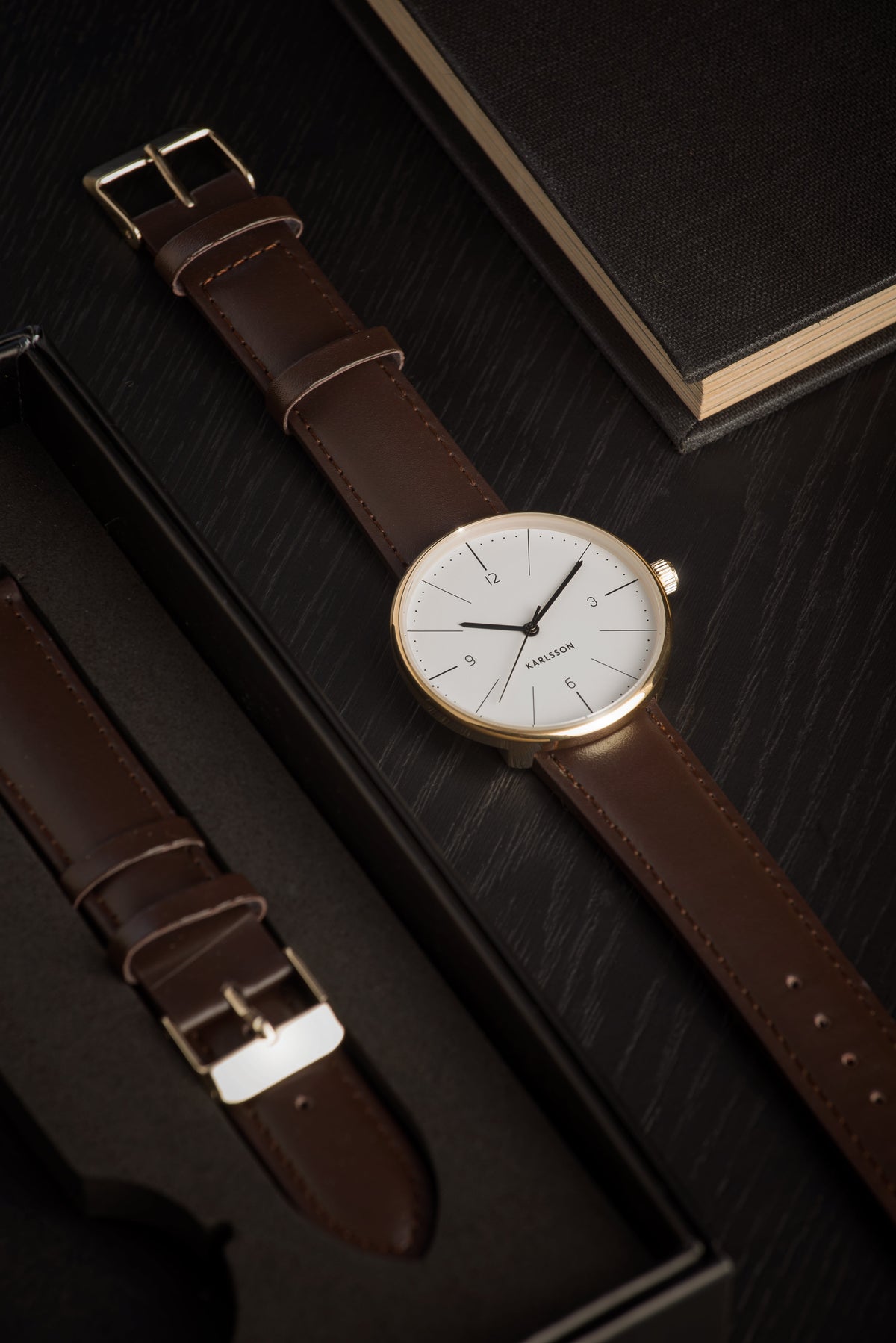 Karlsson Normann watch with a white dial on a dark surface beside a spare strap and a black notebook