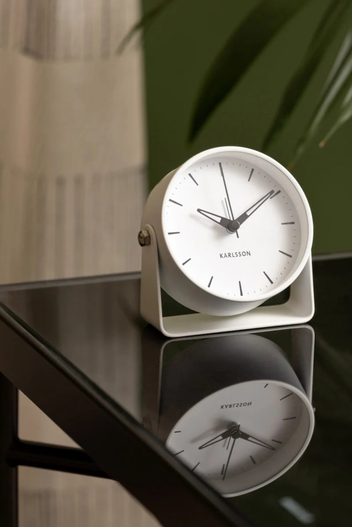 White Karlsson Calm alarm clock on a black glass side table with a plant behind