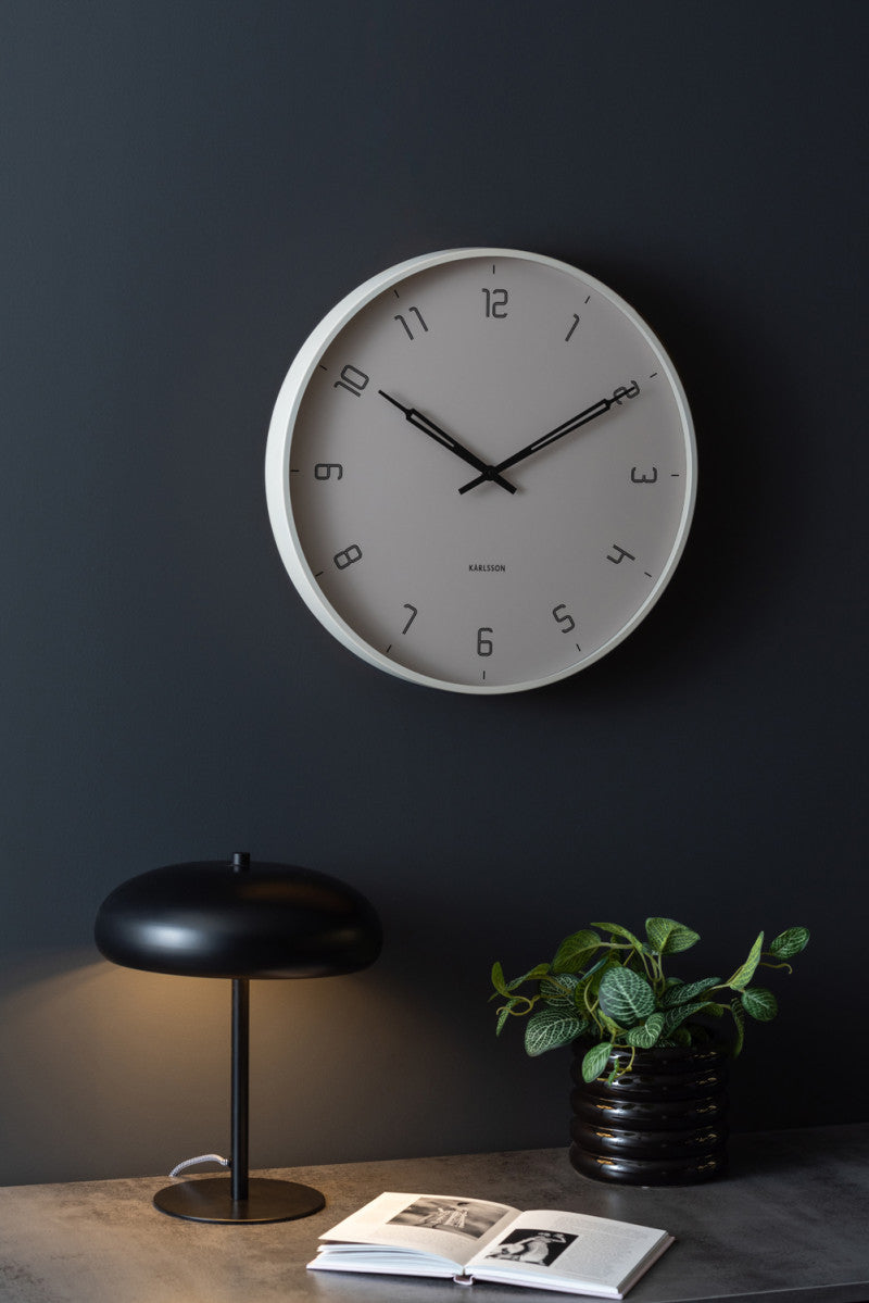Warm grey Stark wall clock on a dark wall above a desk with a lamp and a plant