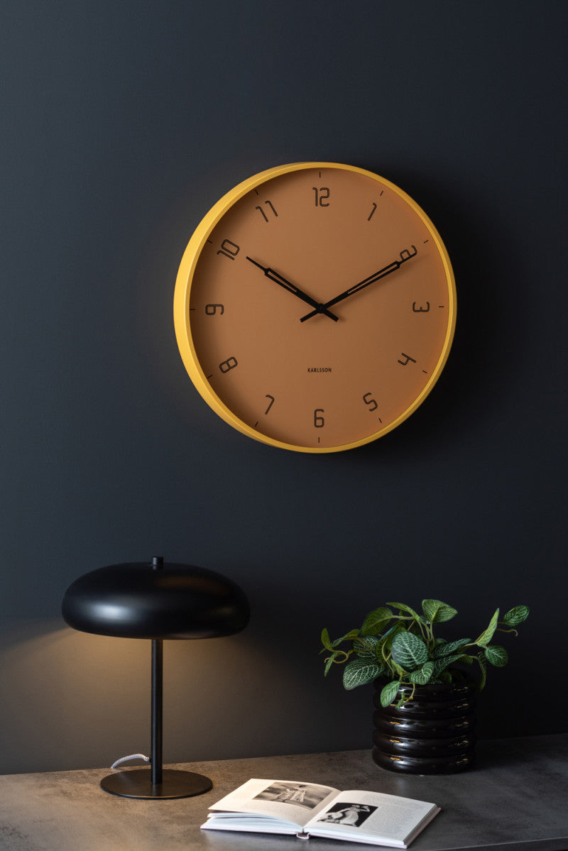 Honey yellow Stark wall clock on a dark wall above a desk with a lamp