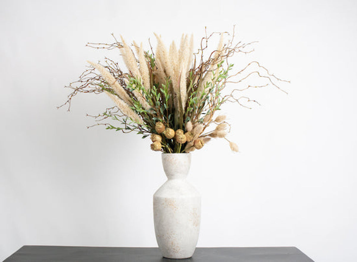 Tall mixed arrangement with fountain grass in a white vase on a dark shelf