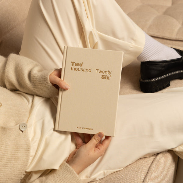 Person holding a beige notebook with 'Two thousand Twenty Six' on the cover, sitting on a couch.