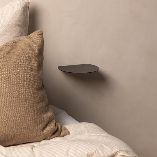 Empty black FOLD Round Shelf on a plaster wall beside a bed