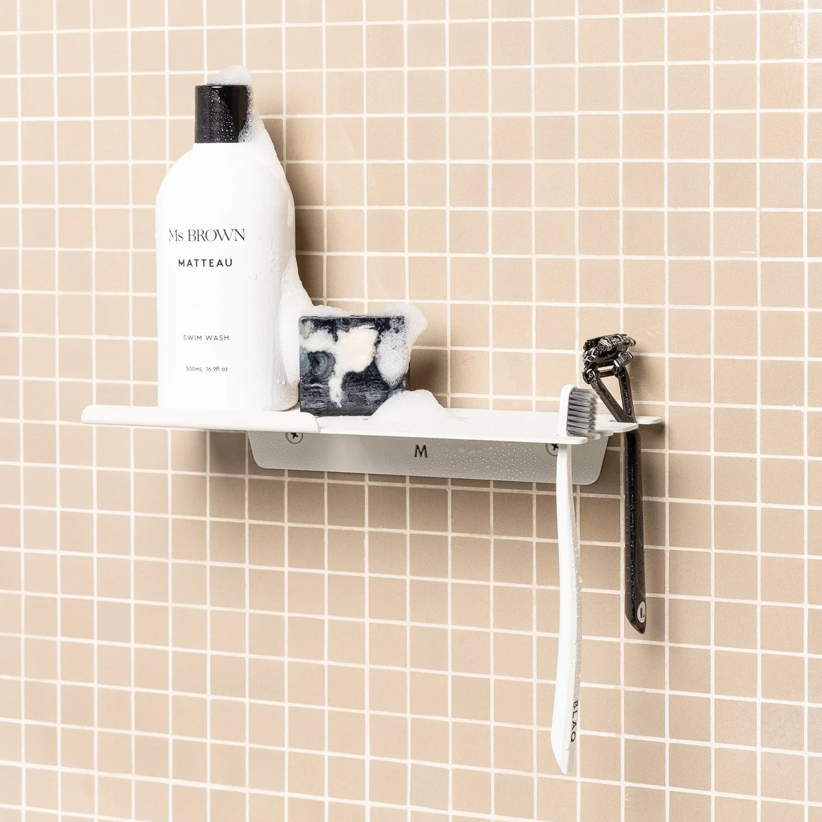 White FOLD shower shelf holding a bottle, razor and toothbrush on pink tiles