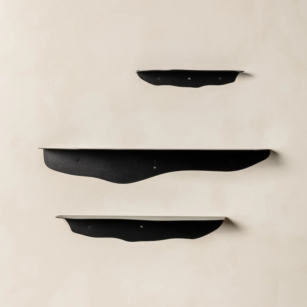 Three empty black wavy shelves staggered on a warm beige plaster wall with soft cast shadows