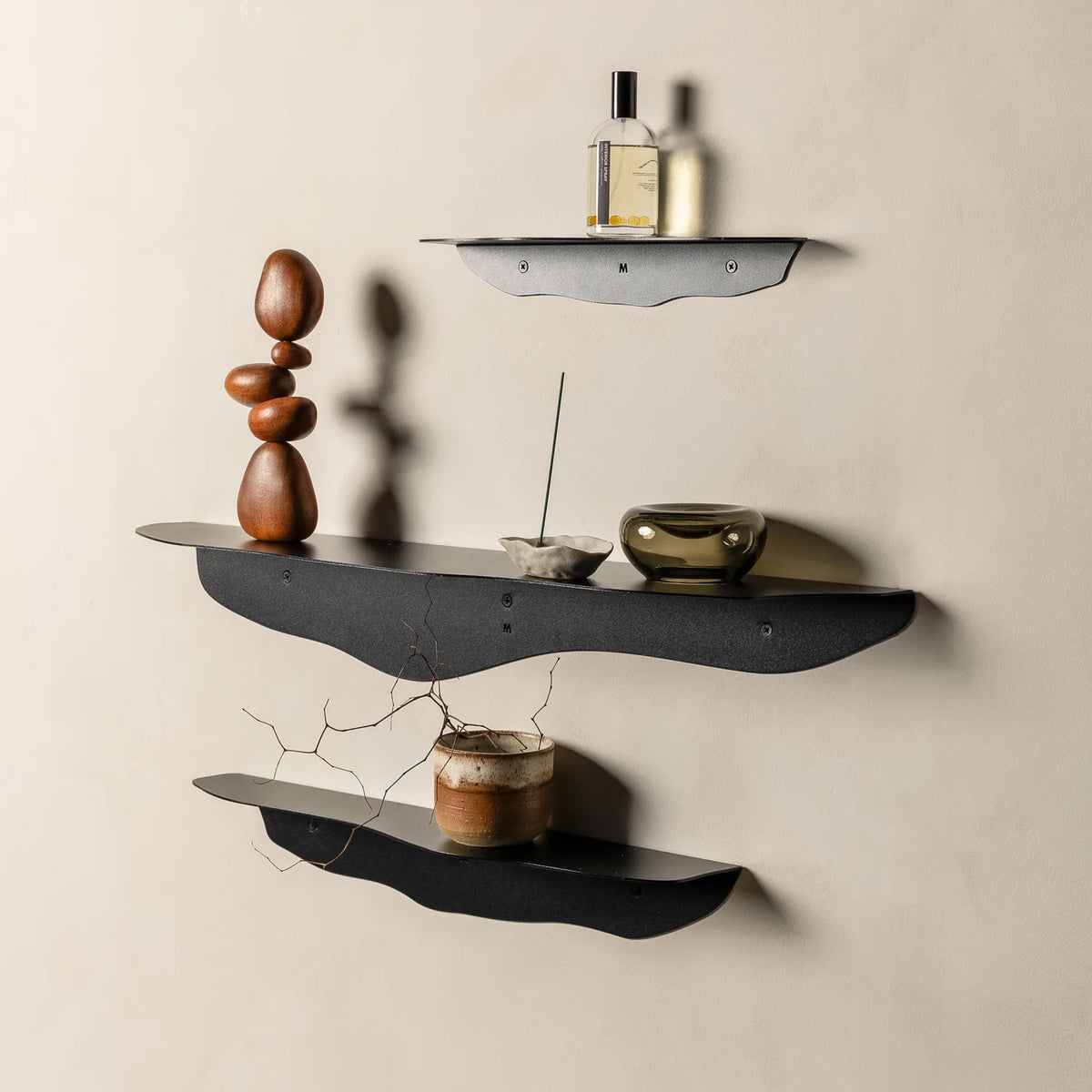 Wider view of three black wavy shelves on beige plaster with glass bottles, brass bowl and candle