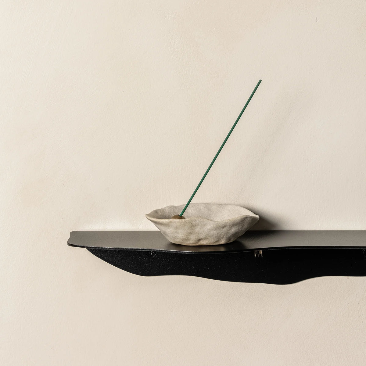 Close crop of a black wavy shelf holding a pale stone pinch dish with a tall green incense stick