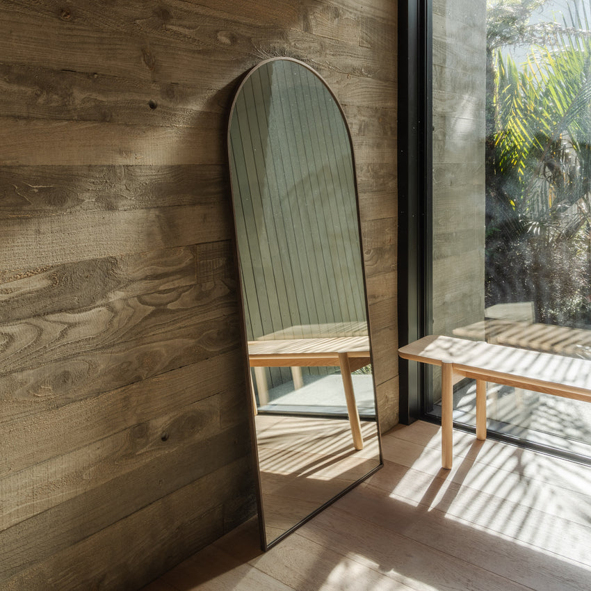 Full length arch mirror with a slim dark frame leaning on a timber plank wall beside glass doors