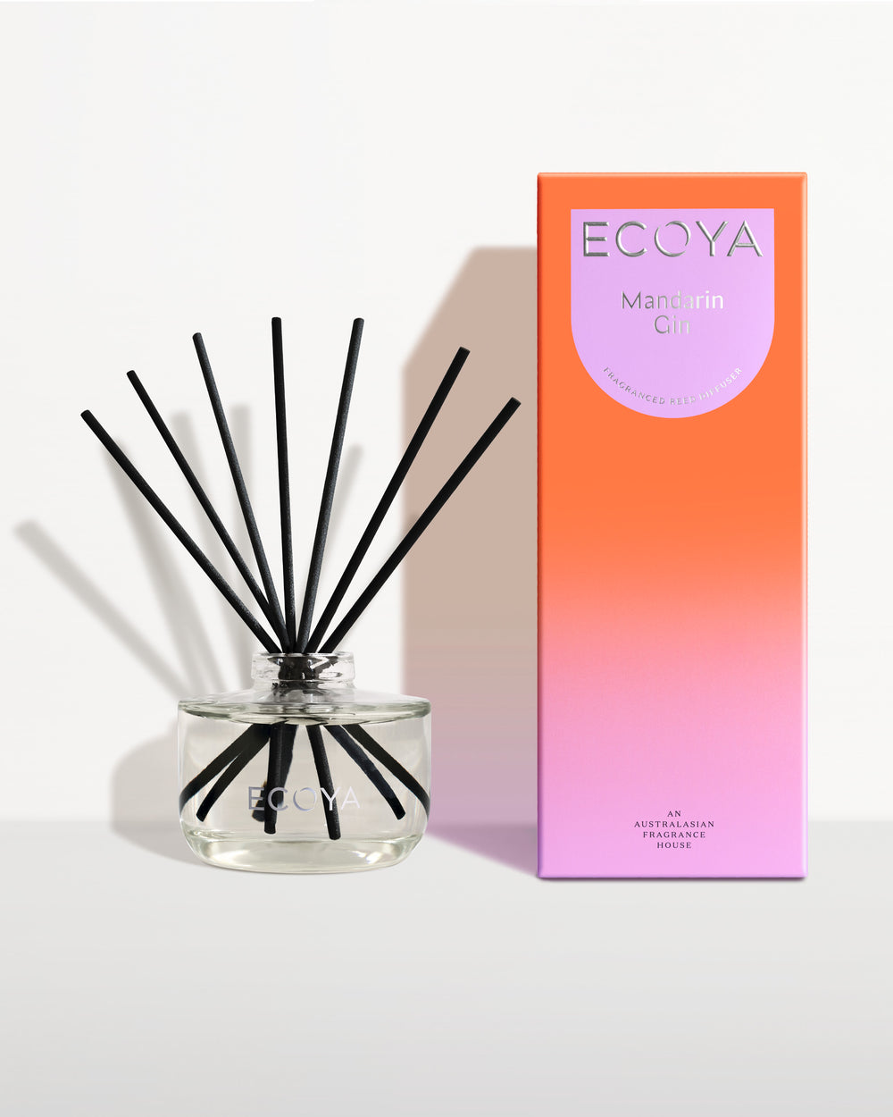 Ecoya Mandarin Gin reed diffuser in a clear glass jar with black reeds, beside its coral gift box