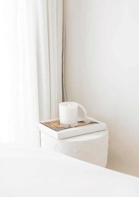 A white soy candle in a ceramic vessel sits atop two books on a textured white side table, positioned beside sheer curtains in a serene, sunlit bedroom corner.

