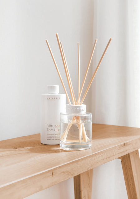 Reed diffuser and Diffuser Top Up bottle on an oak side table against a sheer curtain