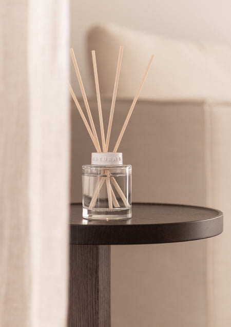 Reed diffuser on a round dark timber side table beside a linen sofa and sheer curtain