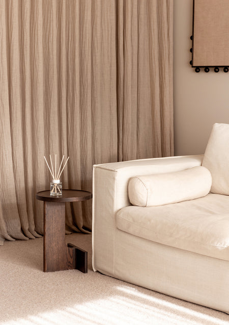 Cream sofa by linen drapes with a small reed diffuser on a dark round side table, portrait view