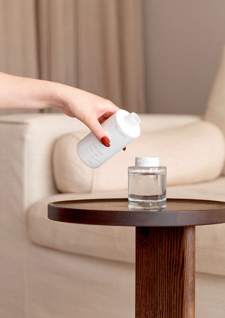 Hand pouring a top up bottle into a reed diffuser on a dark timber side table by a sofa