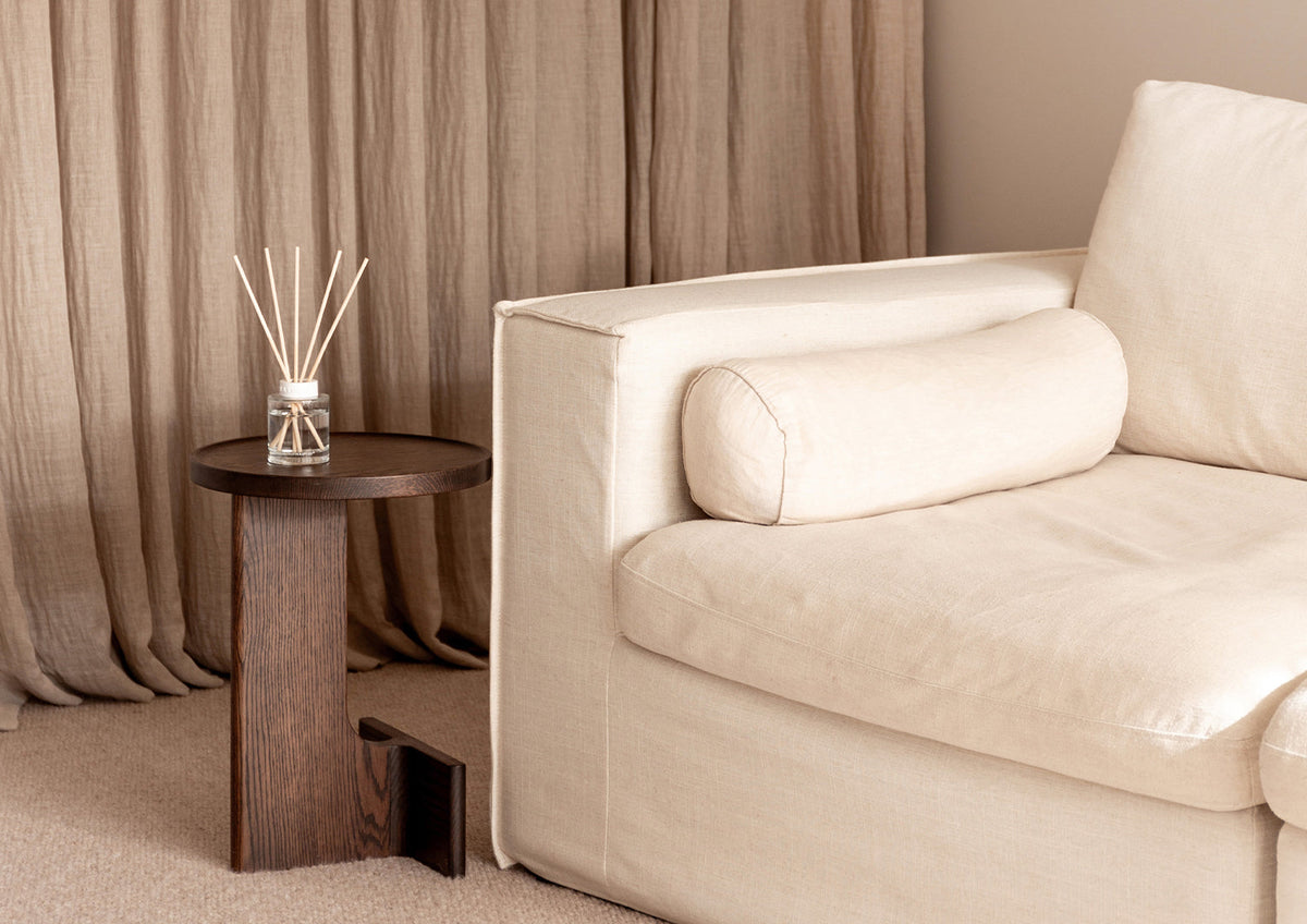 Cream sofa with a bolster cushion beside a dark side table holding a reed diffuser, landscape view