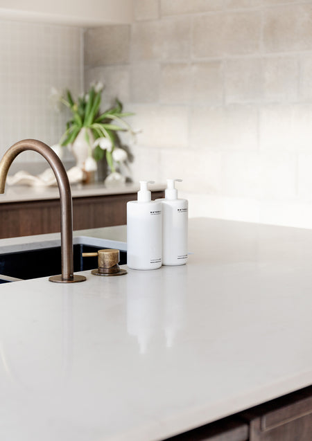 Two Naturals pump bottles beside a bronze gooseneck tap on a white stone kitchen island