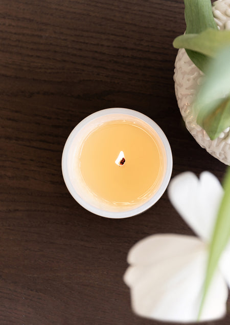 Top view of a lit Naturals candle on dark timber beside a white flower and a green leaf