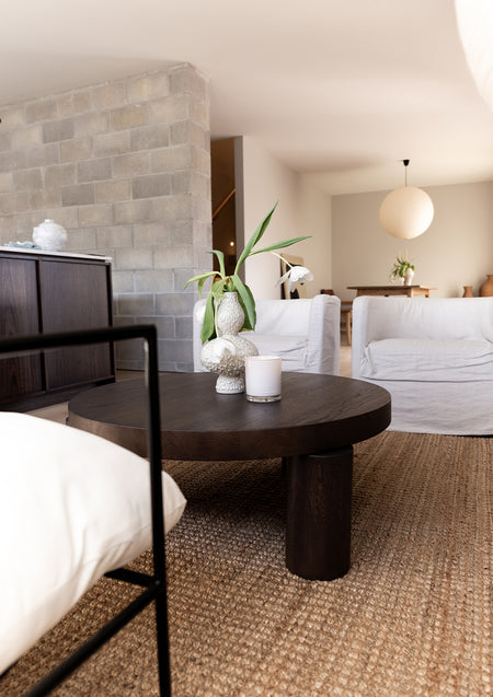 Living room with a dark round coffee table holding a white vase of stems and a lit Naturals candle