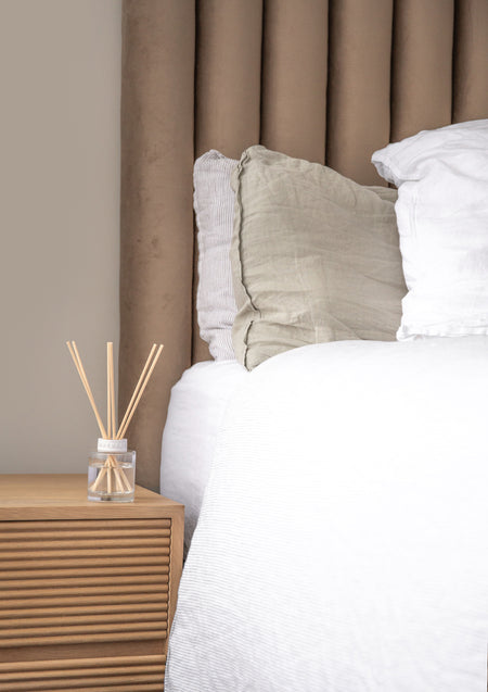 Naturals diffuser on a fluted bedside table beside a made bed with linen cushions
