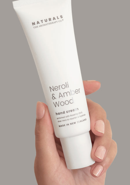 Hand holding the Naturals Neroli & Amber Wood hand cream tube against a grey background