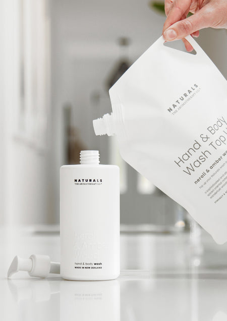 Hand pouring a Hand & Body Wash Top Up pouch into an open Naturals bottle on a white bench