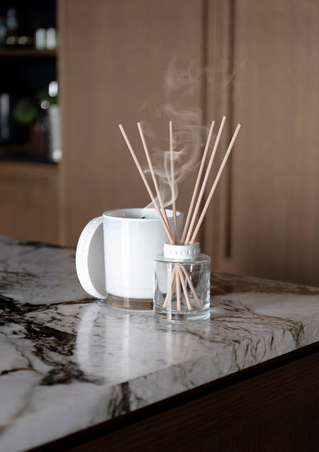 Naturals reed diffuser beside a steaming white mug on a veined marble bench against dark timber