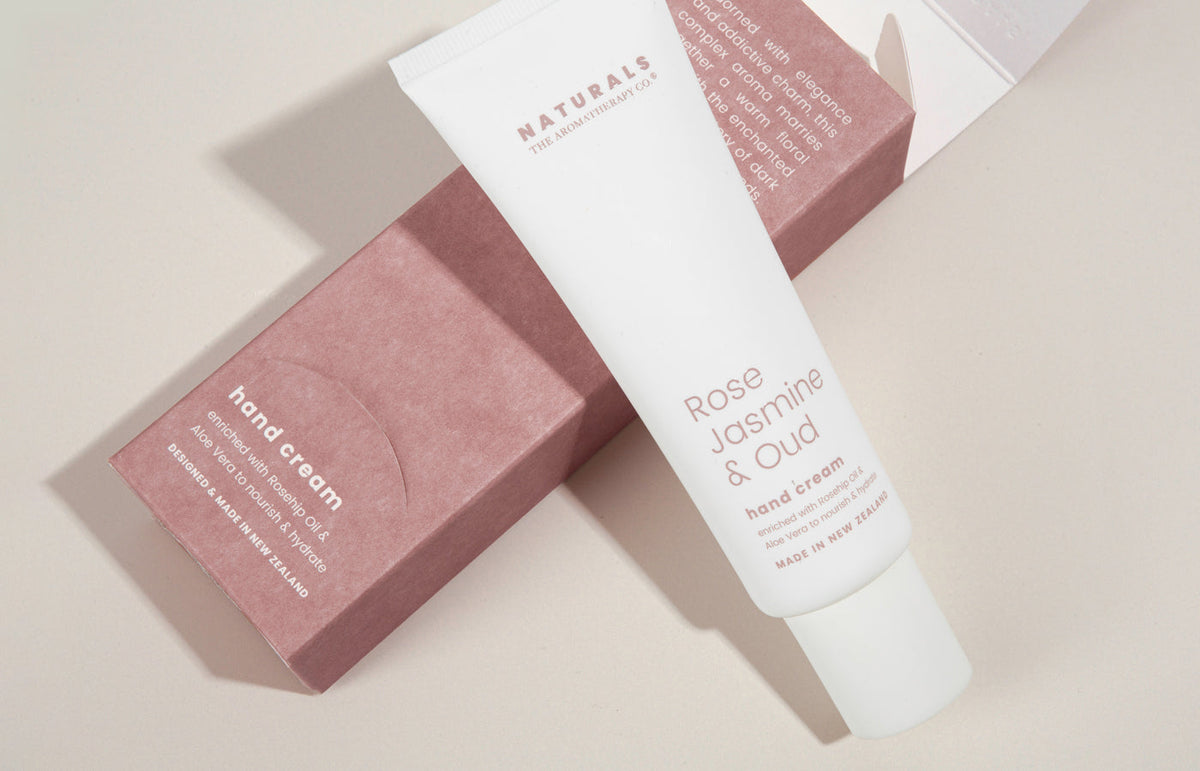 Rose Jasmine & Oud hand cream tube leaning on its pink box on a pale curved surface, landscape view