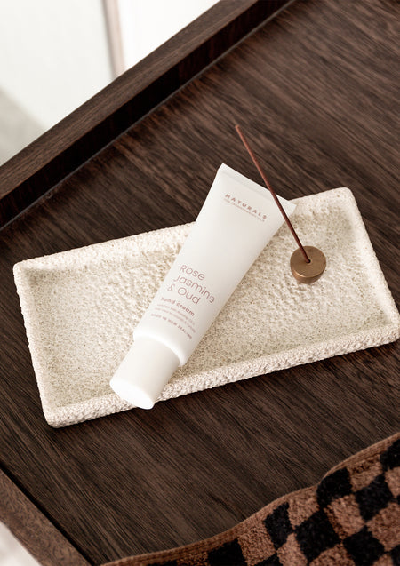 Rose Jasmine & Oud hand cream tube on a textured cream tray with an incense stick on dark timber