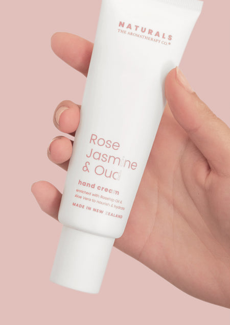 Hand holding the Naturals Rose Jasmine & Oud hand cream tube against a dusty pink background