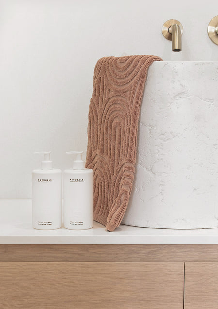 Two Naturals pump bottles on a white vanity beside a stone basin draped with a rust towel