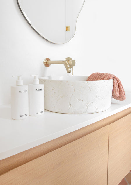 Two Naturals pump bottles beside a textured white stone basin and brass tap on an oak vanity