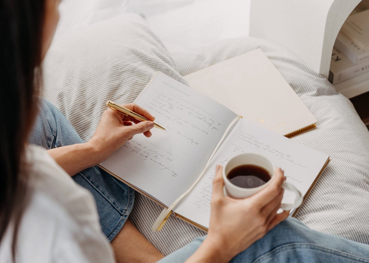 A woman writing in the YOU. journal on a bed while holding a cup of coffee