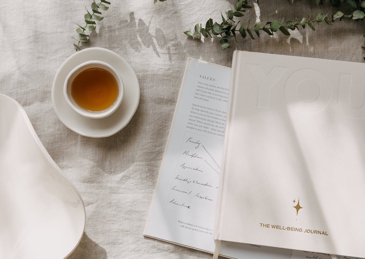 The cream YOU. journal open beside a handwritten page and a cup of tea on linen, with eucalyptus stems alongside