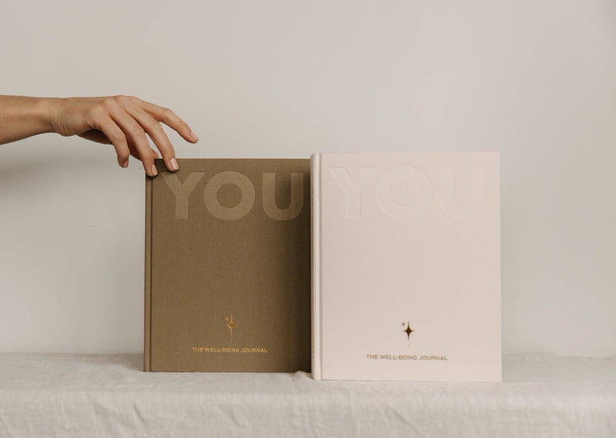 A hand reaching for the sand and cream editions of the YOU. journal standing side by side on a linen covered table