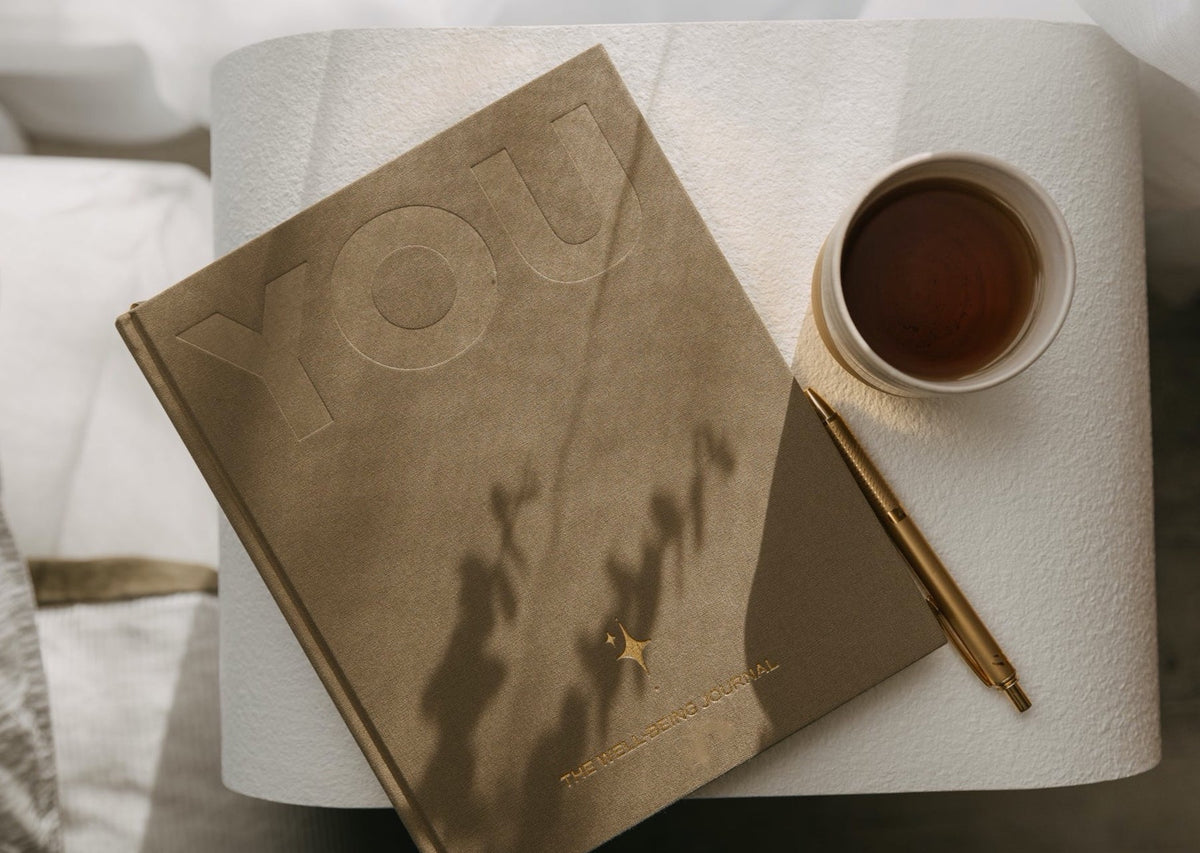 The sand coloured YOU. journal on a round side table with a gold pen and a cup of tea, in dappled light