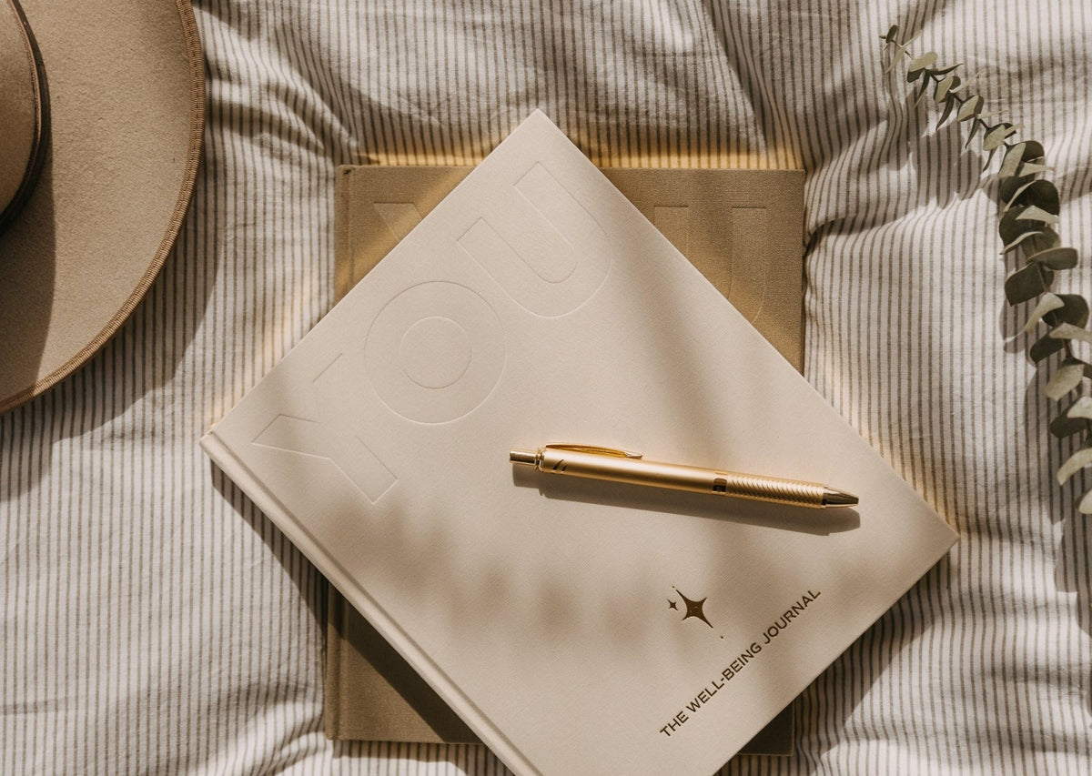 The cream YOU. journal with a gold pen on striped bedding, styled with a felt hat and eucalyptus stems