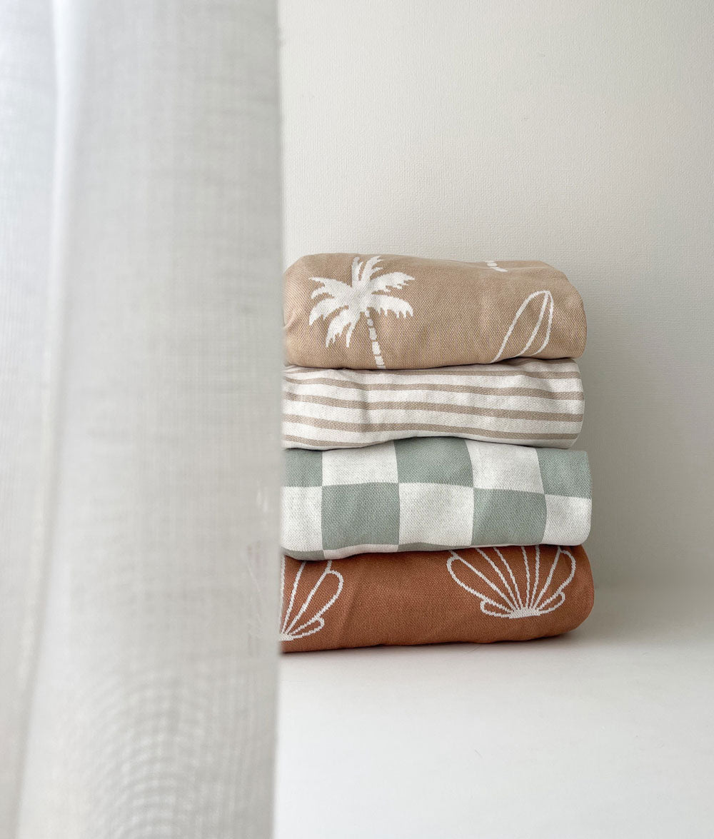 Stack of folded jersey fitted sheets in Surfing Palm, stripe, sage gingham and shell prints by a sheer curtain