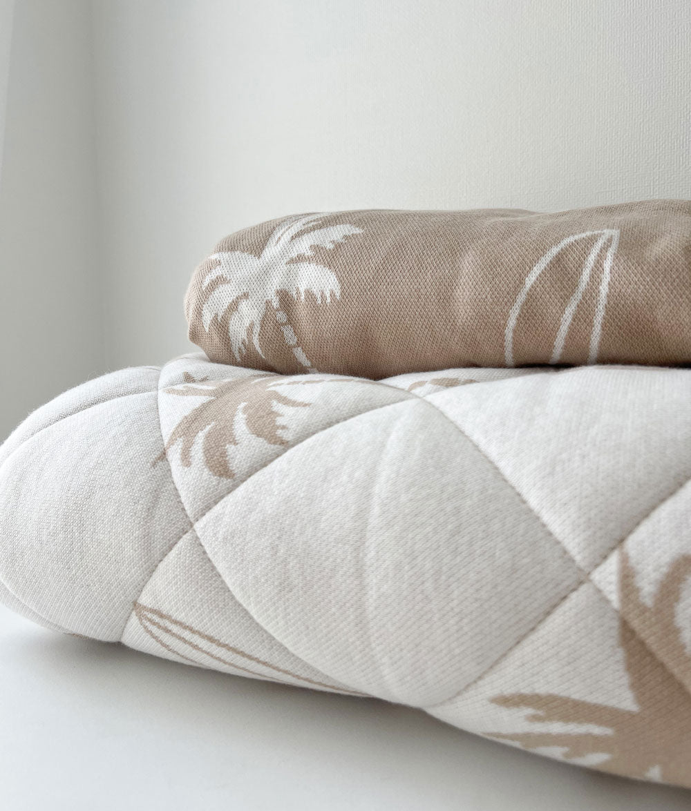 Close-up of a folded natural Surfing Palm reversible quilt topped with a sand Surfing Palm fitted sheet