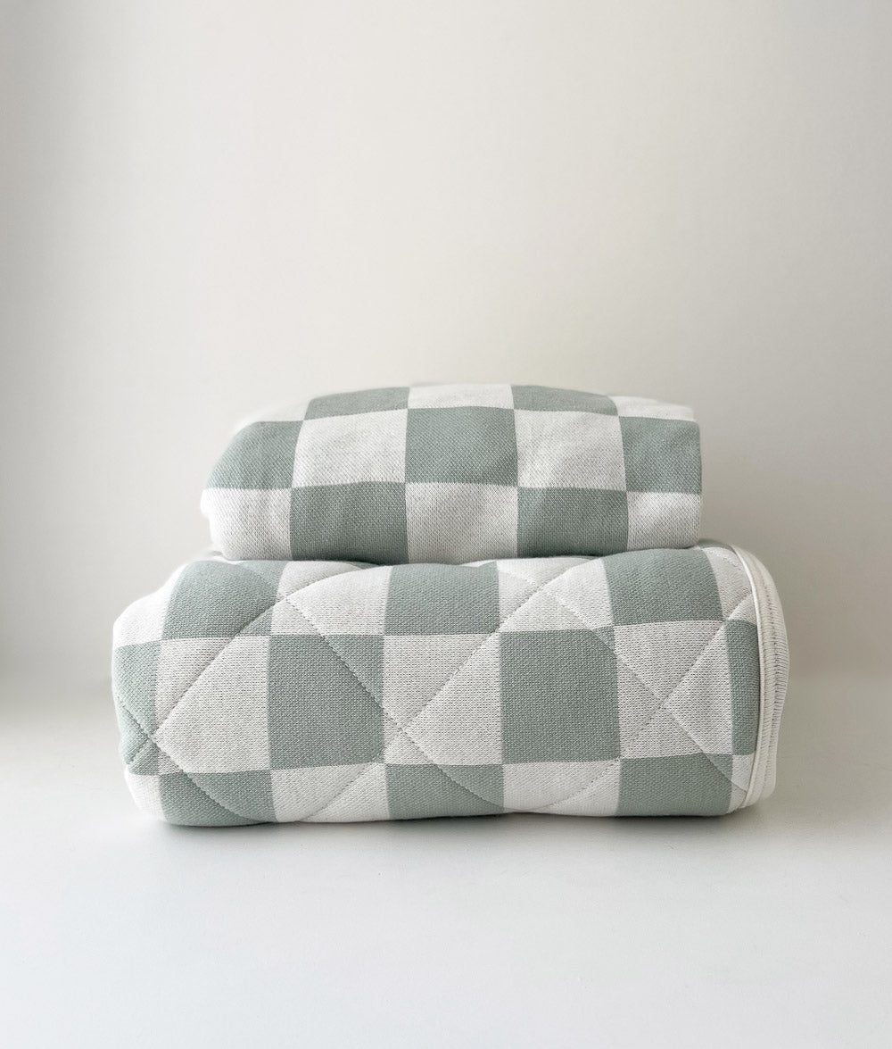 Folded Sage Gingham jersey fitted sheet on top of a rolled Sage Gingham reversible quilt