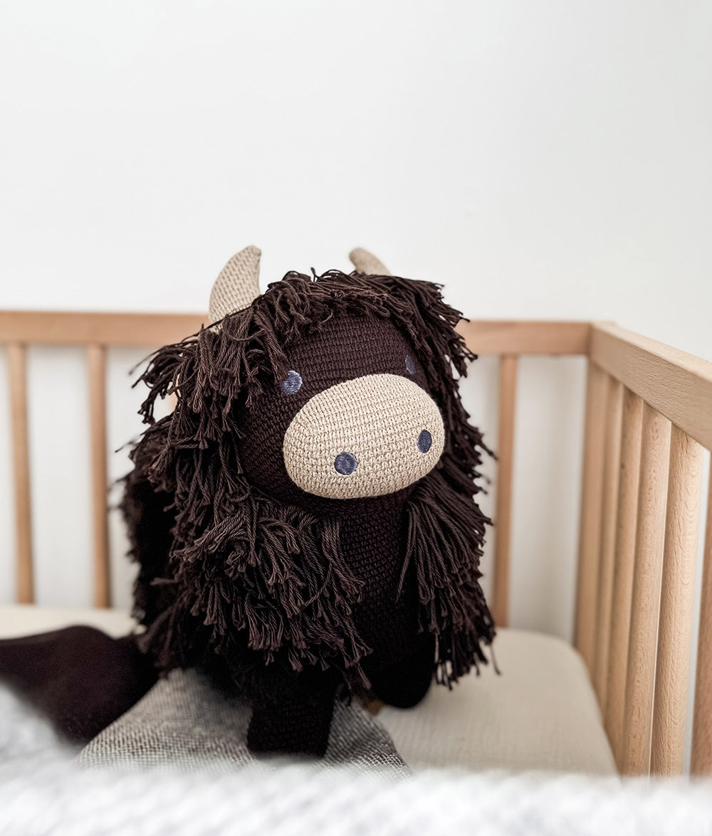 Fergus the Highland cow soft toy standing in a timber cot, showing his cream horns and muzzle