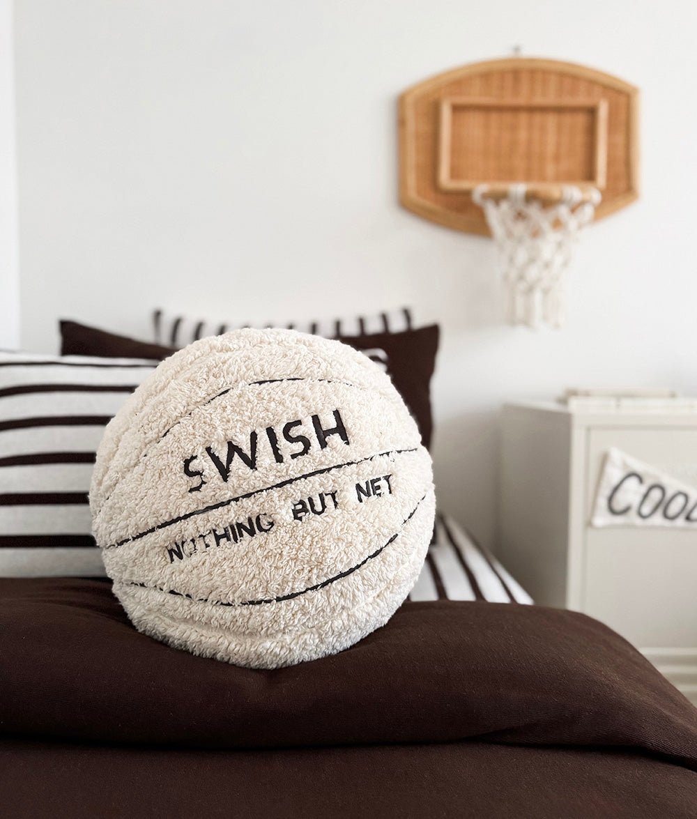 Basketball sherpa cushion on a chocolate duvet with striped pillows and a rattan hoop above