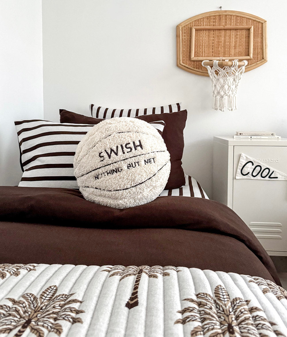 Basketball sherpa cushion propped on a chocolate duvet with a palm print quilt across the foot