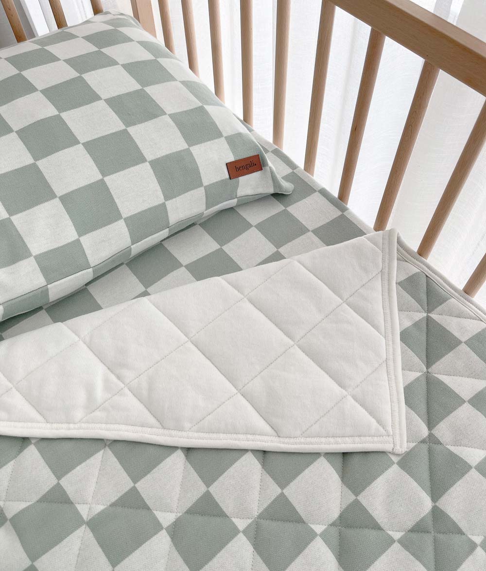 Sage Gingham cot quilt folded back to its ivory side beneath a matching pillowcase in a timber cot