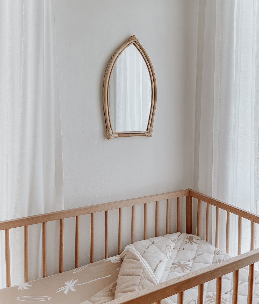 Timber cot with a folded Surfing Palm reversible cot quilt beneath a surfboard-shaped rattan mirror