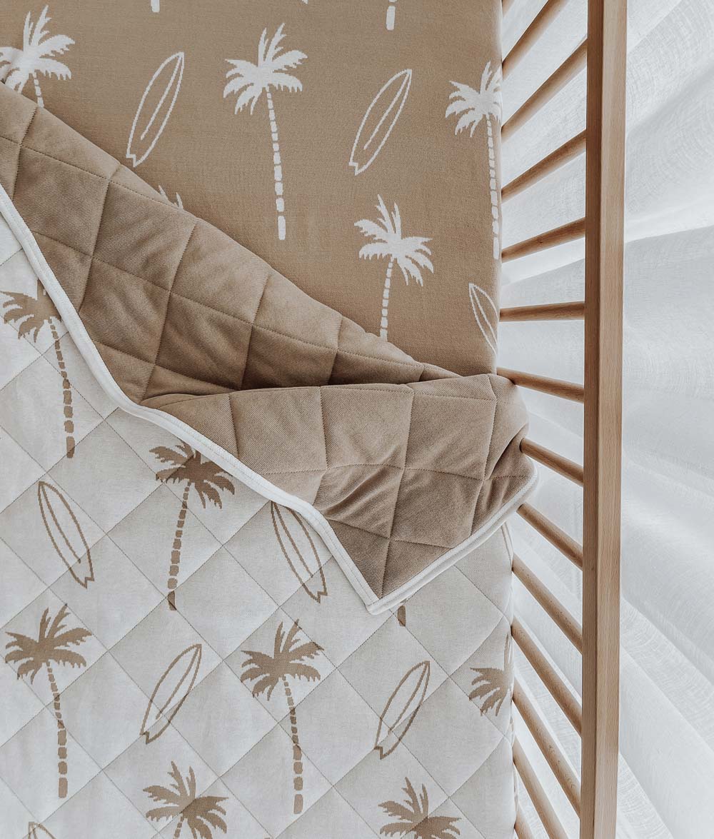 Surfing Palm reversible quilt draped over a timber cot rail, showing the palm print and plain sand reverse