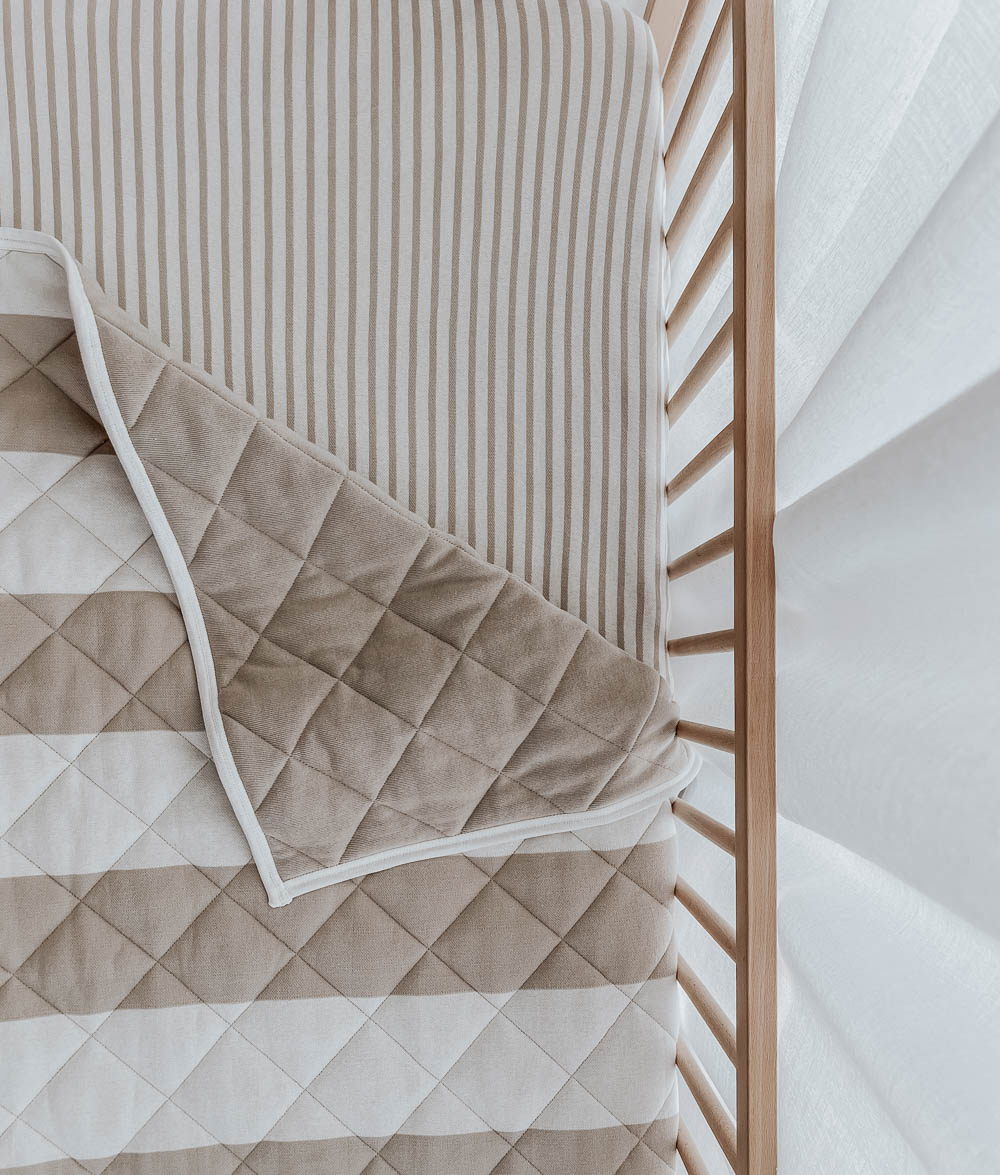 Overhead view of a greige stripe reversible quilt turned back on a greige pinstripe cot sheet