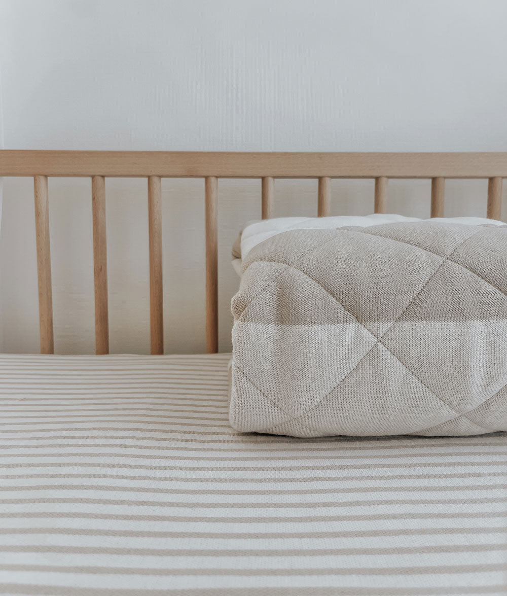 Rolled greige stripe quilt resting on a greige pinstripe jersey cot sheet in a timber cot
